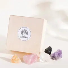 Drewniane pudełko z naklejką "BASIC CRYSTALBOX" i pięcioma surowymi kamieniami: bursztynowym, różowym kwarcem, kryształem górskim, czarnym turmalinem i ametystem. Każdy cenny kamień emanuje naturalnym blaskiem.