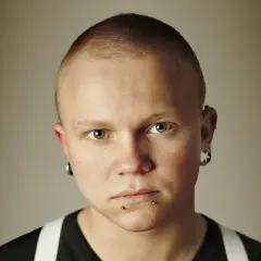 Mężczyzna z niebieskimi oczami i kolczykiem w nosie. Ma krótko ostrzyżone włosy i kolczyki w uszach.