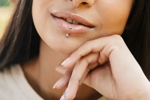 Usta z błyszczącym błyszczykiem i subtelnym center labret. Dłoń podtrzymuje podbródek, podkreślając delikatny uśmiech.