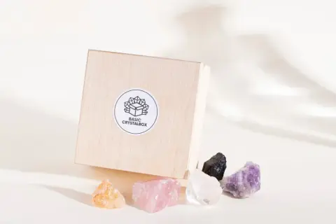 Drewniane pudełko z naklejką "BASIC CRYSTALBOX" i pięcioma surowymi kamieniami: bursztynowym, różowym kwarcem, kryształem górskim, czarnym turmalinem i ametystem. Każdy cenny kamień emanuje naturalnym blaskiem.