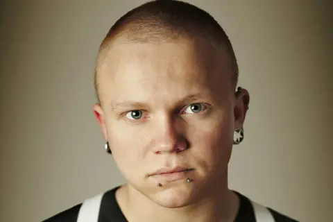 Mężczyzna z niebieskimi oczami i kolczykiem w nosie. Ma krótko ostrzyżone włosy i kolczyki w uszach.