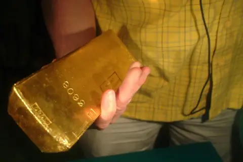 Dłoń trzyma błyszczącą sztabkę złota 5kg z wybitym numerem seryjnym.