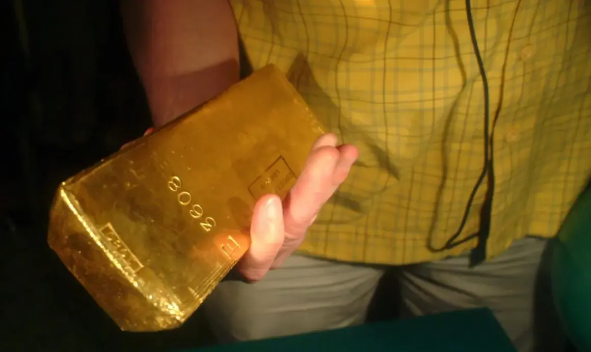 Dłoń trzyma błyszczącą sztabkę złota 5kg z wybitym numerem seryjnym.