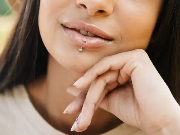 Usta z błyszczącym błyszczykiem i subtelnym center labret. Dłoń podtrzymuje podbródek, podkreślając delikatny uśmiech.