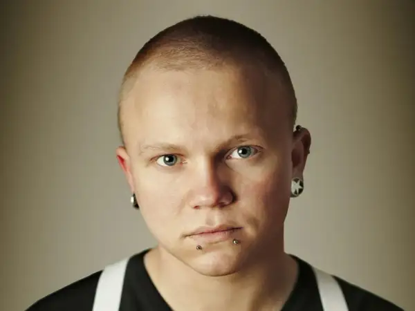 Mężczyzna z niebieskimi oczami i kolczykiem w nosie. Ma krótko ostrzyżone włosy i kolczyki w uszach.